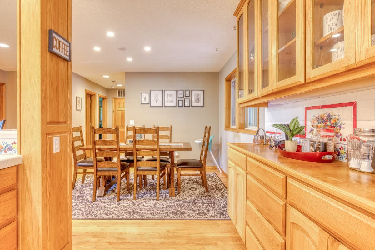 Cozy dining area with a wooden table and six chairs, set on a patterned area rug. Light wood cabinetry with glass fronts and a coffee station are visible in the background, along with framed artwork on the wall.