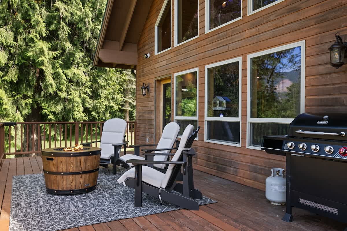 A wooden outdoor deck furnished with black Adirondack chairs, a barrel-style fire pit, and a black gas grill, all resting on a patterned outdoor rug, with a view of the wooden house exterior and surrounding trees.