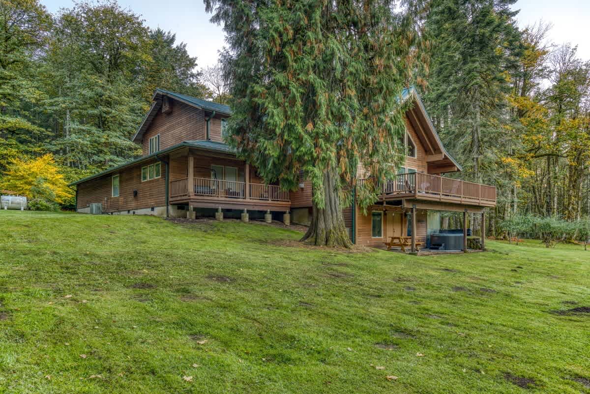 A wide view of the wooden lodge's side and back, showcasing its multi-level structure with decks, nestled on a grassy slope amidst dense evergreen and deciduous trees.