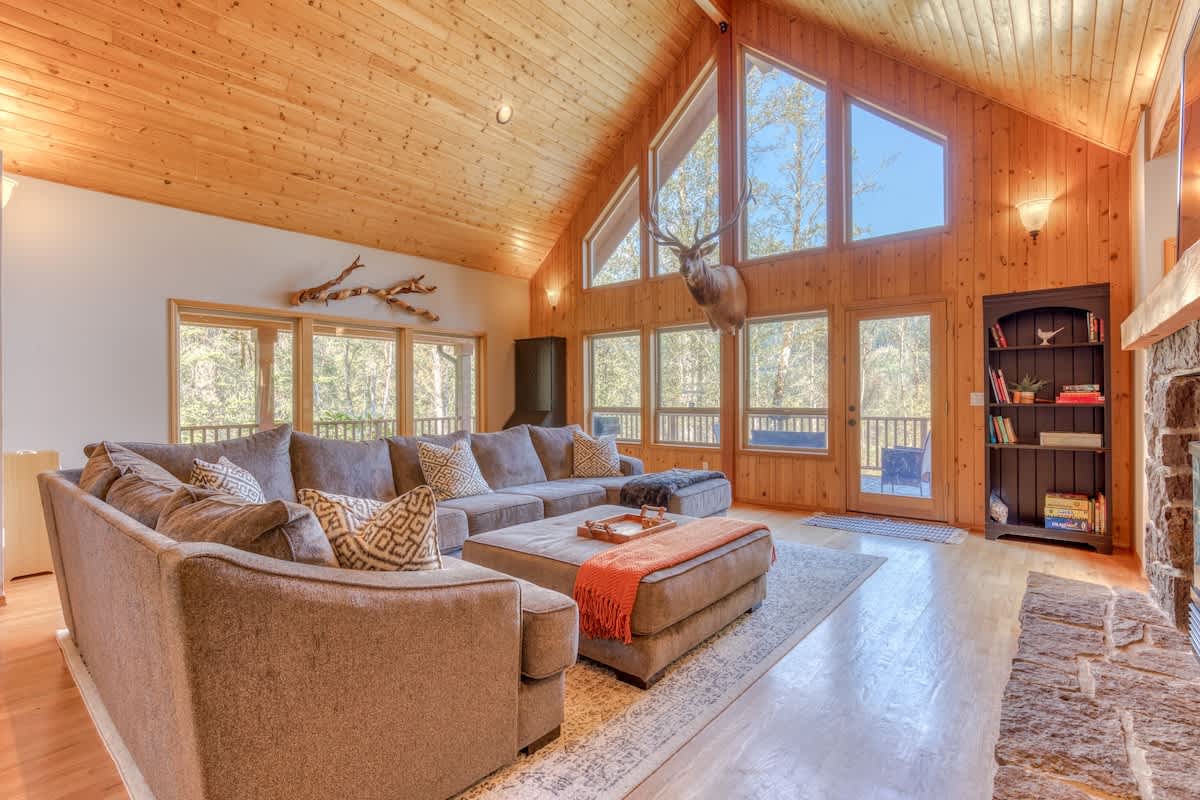 A spacious living room with a large gray sectional sofa, an orange throw blanket on an ottoman, a stone fireplace, and expansive windows offering views of trees. The room features a vaulted wooden ceiling and light hardwood floors.