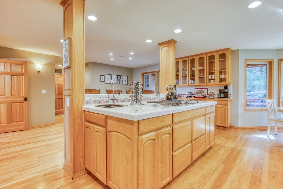 Open-concept kitchen featuring a large island with a gas cooktop and white tiled countertop, surrounded by light wood cabinetry and hardwood floors. A breakfast nook with a round white table is visible through the bay windows in the background.
