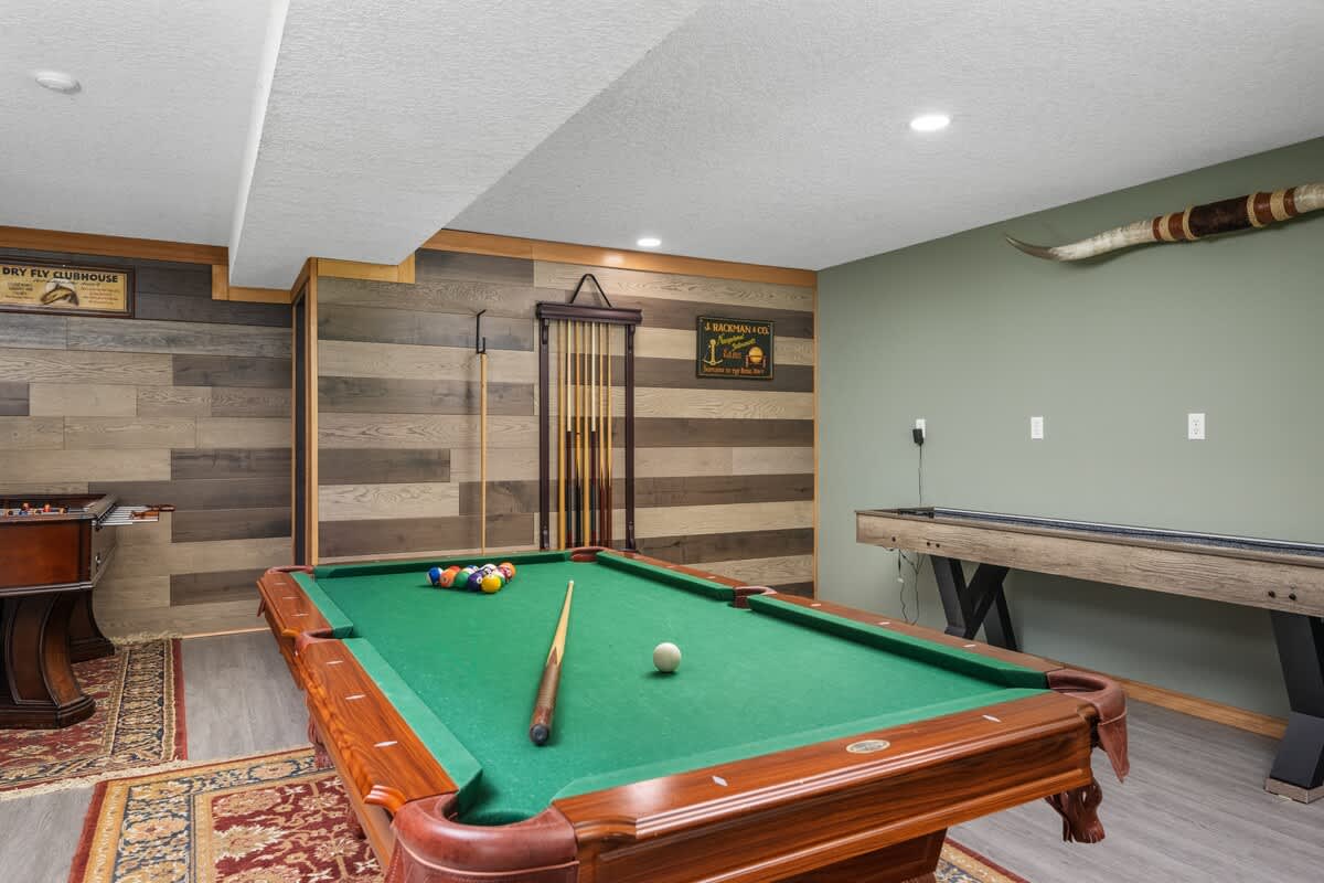 A spacious game room featuring a green-felted pool table with cues and balls, a long shuffleboard table, and a wall with horizontal wood paneling and a sage green painted section.