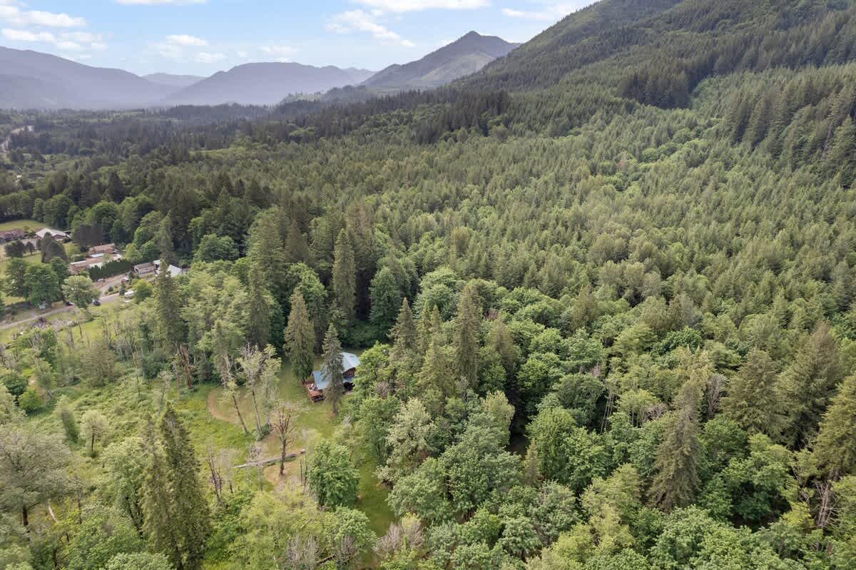 A high aerial view reveals the property nestled within an expansive, lush green forest, with distant mountains under a partly cloudy sky, highlighting the secluded natural setting.