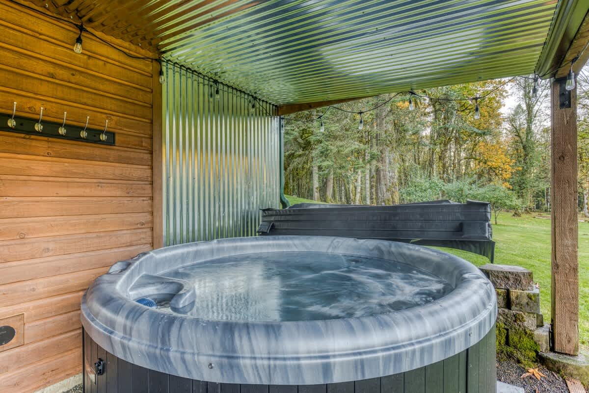A bubbling hot tub with a grey and black marbled finish is situated under a corrugated metal roof, next to a wooden wall, with string lights overhead and a lush green forest in the background.