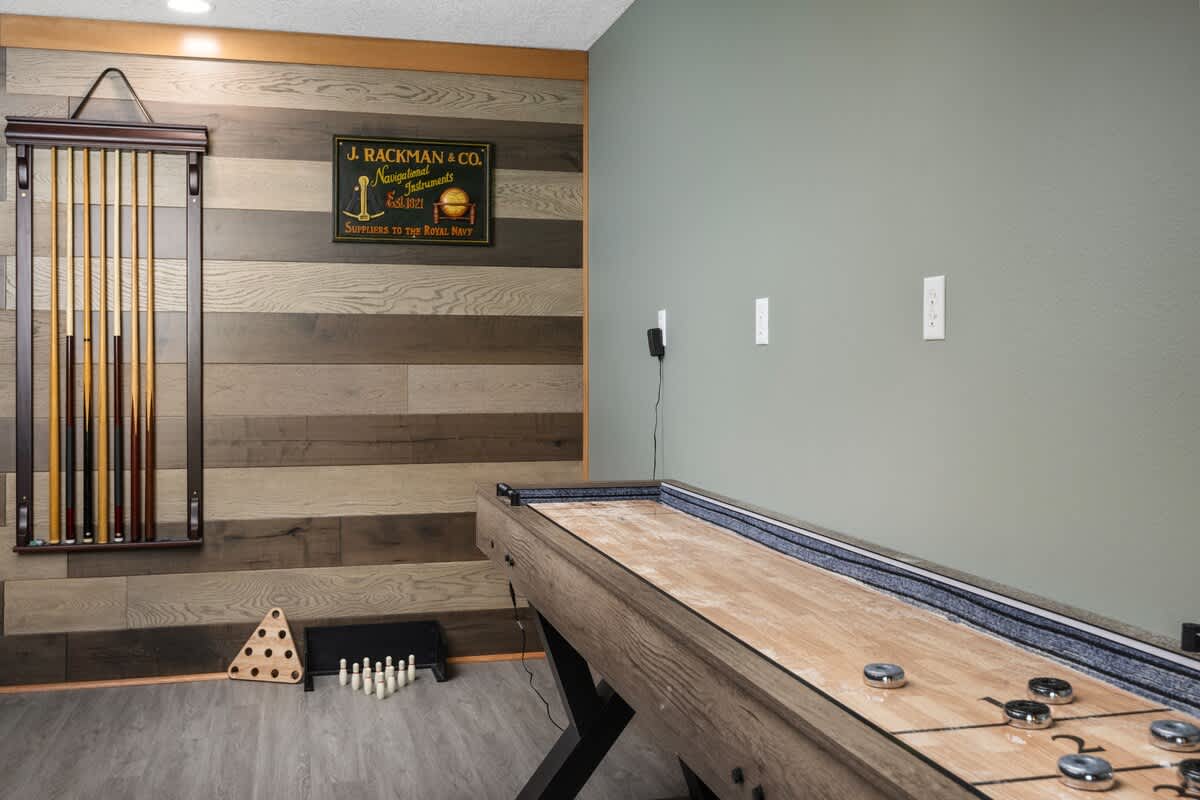 A detailed view of a rustic wooden shuffleboard table with pucks, alongside a wall-mounted cue rack holding several pool cues, set against a backdrop of horizontal wood paneling and a sage green wall.