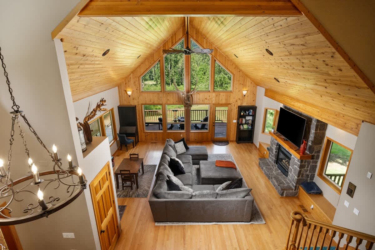 An aerial view of a spacious living room with a large gray sectional sofa, a small dining table, a stone fireplace, and a TV. The room features a high vaulted wooden ceiling, light hardwood floors, and large windows overlooking trees.
