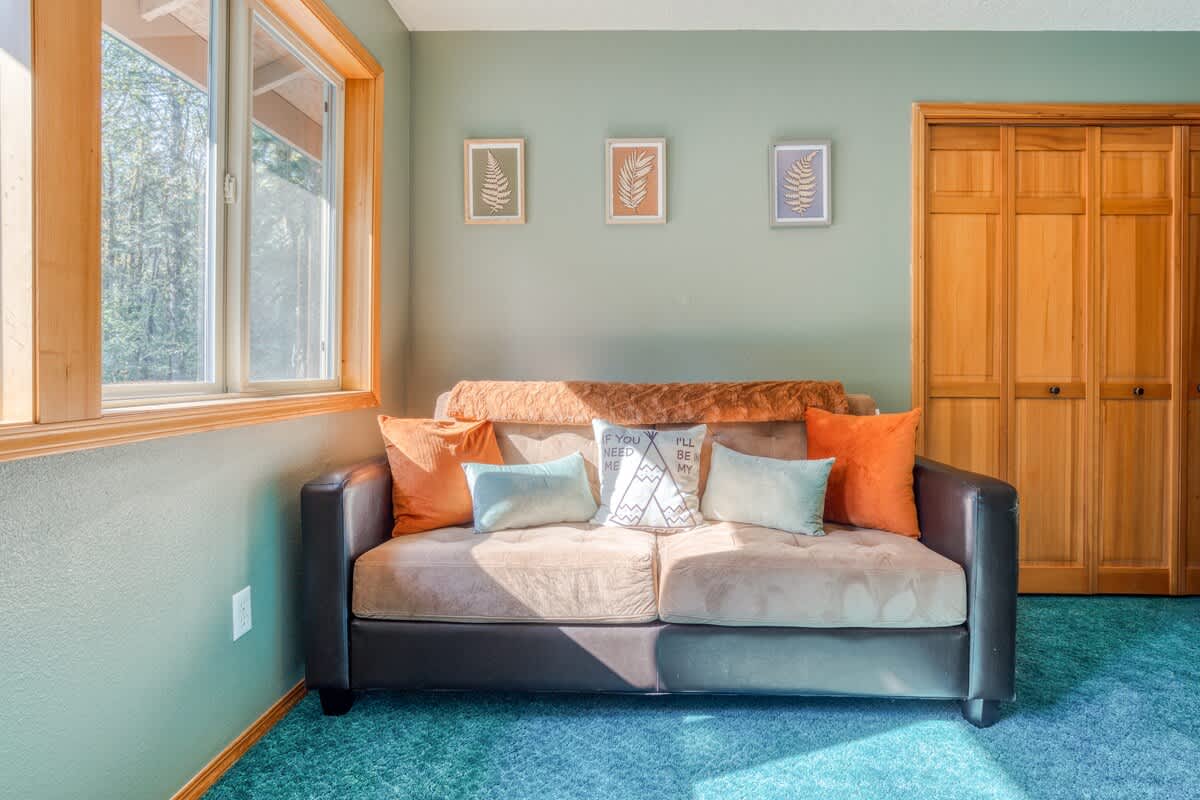 A comfortable brown sofa with various throw pillows sits against an olive green wall, beneath three framed botanical prints, with large windows offering a view of greenery.