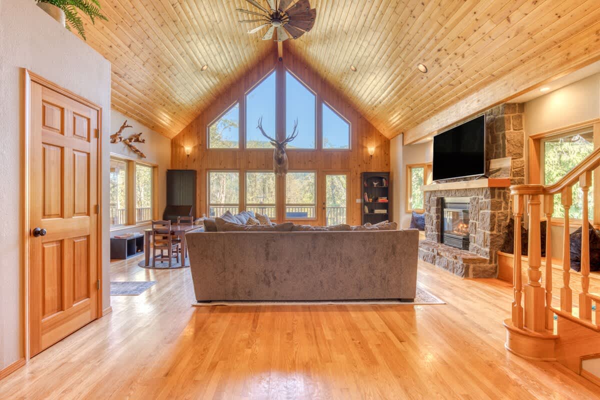 A grand living room boasts a vaulted wooden ceiling, a large sectional sofa, a stone fireplace, and expansive windows offering panoramic views of the surrounding nature, with a mounted deer head as a focal point.