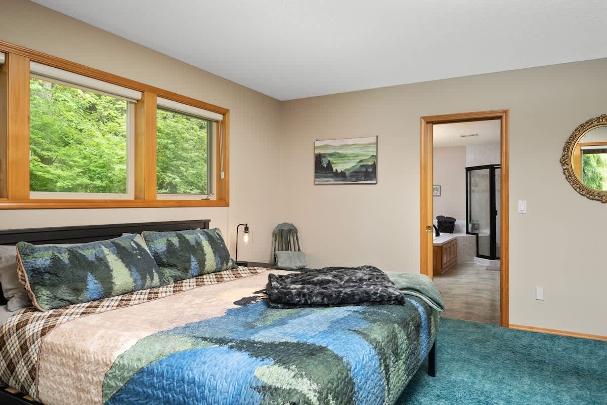 A view of the bedroom showing the bed with its patterned comforter, large windows, and an open doorway leading to an ensuite bathroom. The bathroom features a large tub and a glass-enclosed shower.