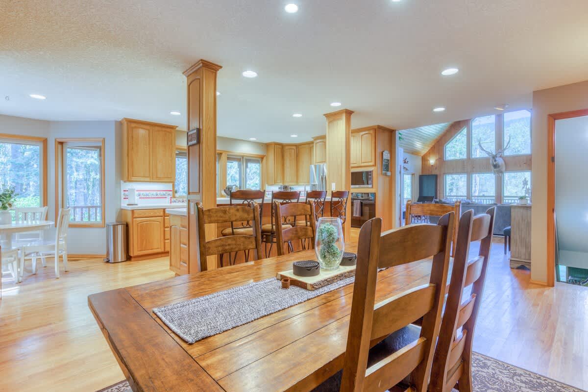 A spacious kitchen and dining area featuring a large wooden dining table with chairs in the foreground, and a kitchen with light wood cabinetry and a breakfast bar in the background. Light hardwood floors connect the spaces.