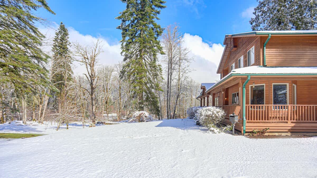 Winter view of the wooden lodge, showcasing its side with a covered porch and a snow-covered lawn, surrounded by tall evergreen and bare deciduous trees under a bright blue sky.