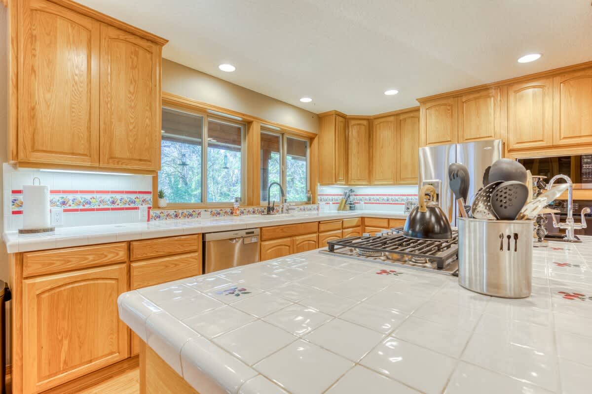 Detailed view of the kitchen island with a gas cooktop and white tiled countertop, complemented by light wood cabinetry. A stainless steel dishwasher is visible, along with a decorative tiled backsplash featuring red accents.