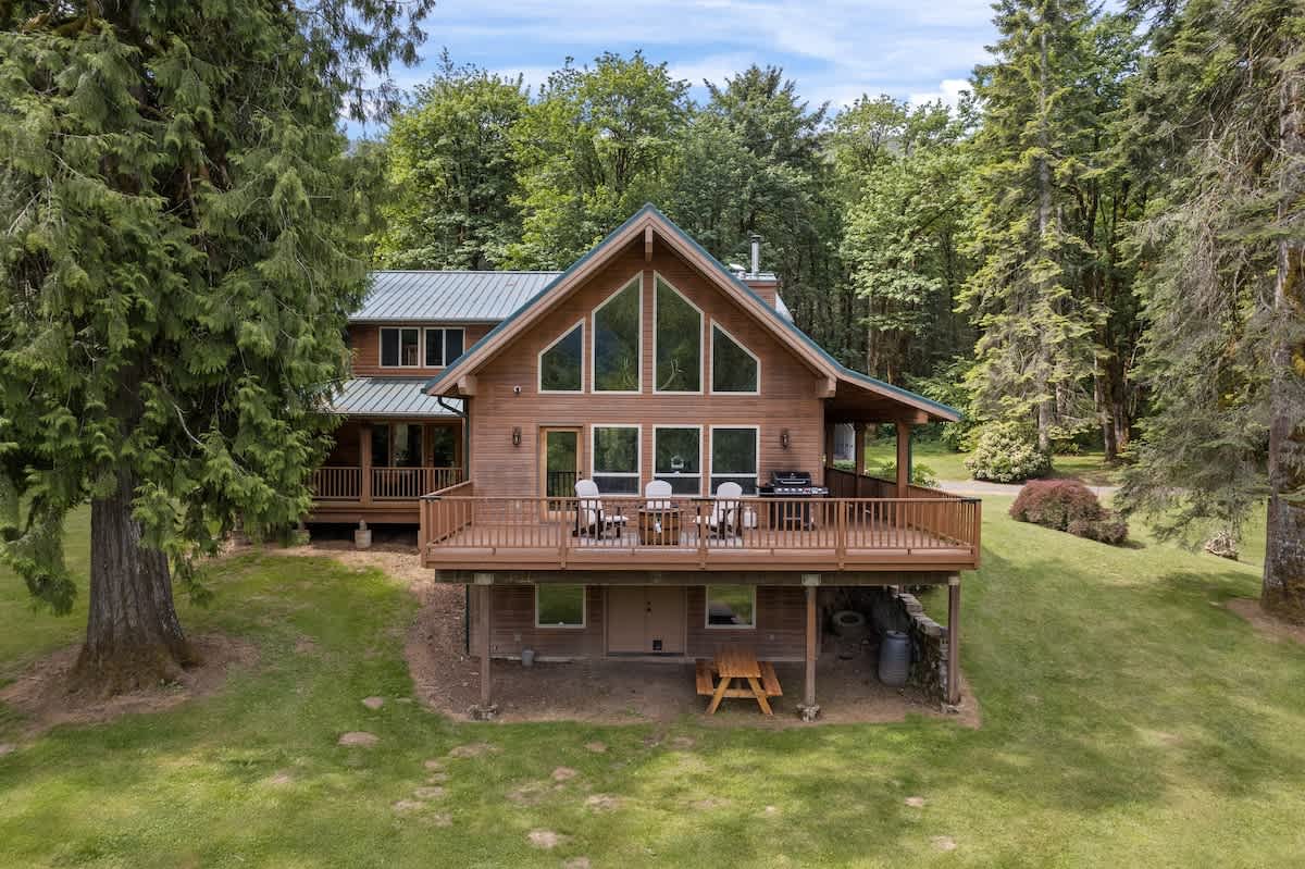 A large, rustic wooden lodge-style house with a prominent A-frame section, multiple windows, and a spacious elevated deck with outdoor seating, surrounded by lush green trees and a manicured lawn.