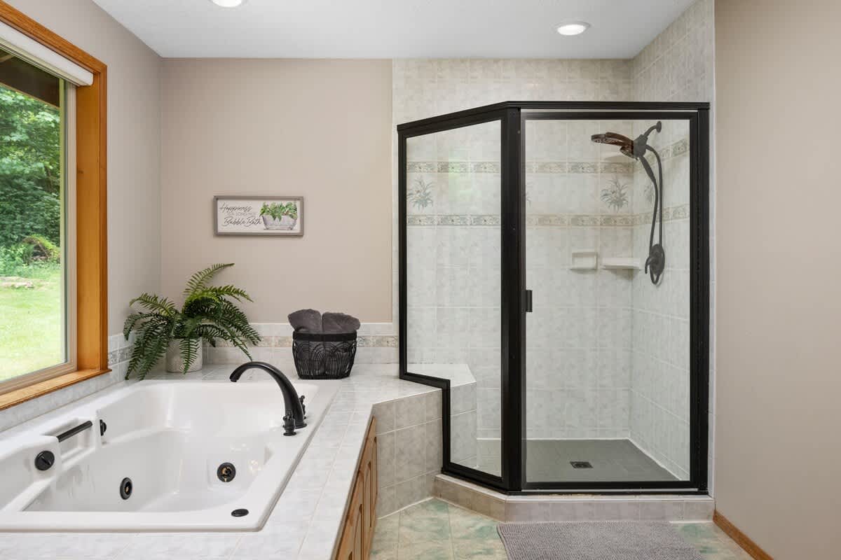 A bright bathroom featuring a white built-in jetted tub with black fixtures, adjacent to a corner shower with black-framed glass doors and light tiled walls. A large window overlooks lush greenery.