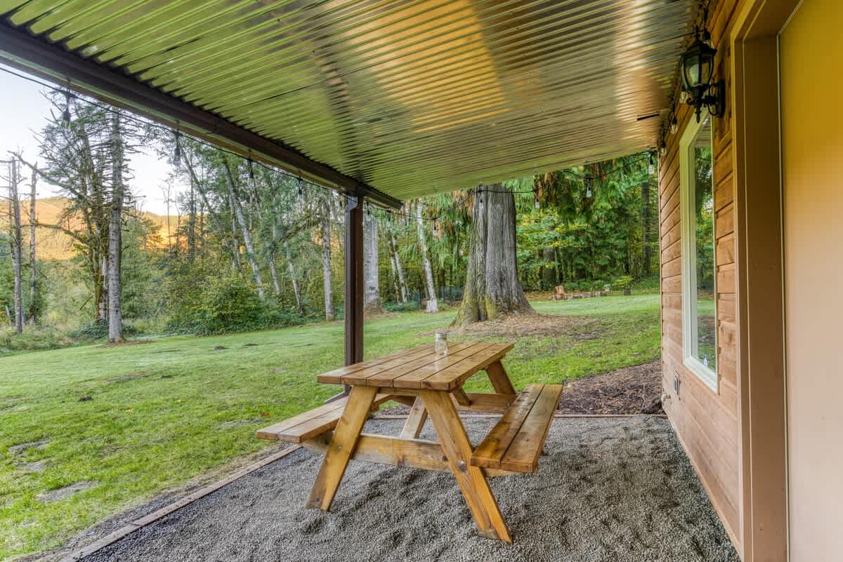A wooden picnic table with benches placed on a gravel ground under a corrugated metal roof, adjacent to the wooden exterior of the house, with a lush green lawn and trees in the background.