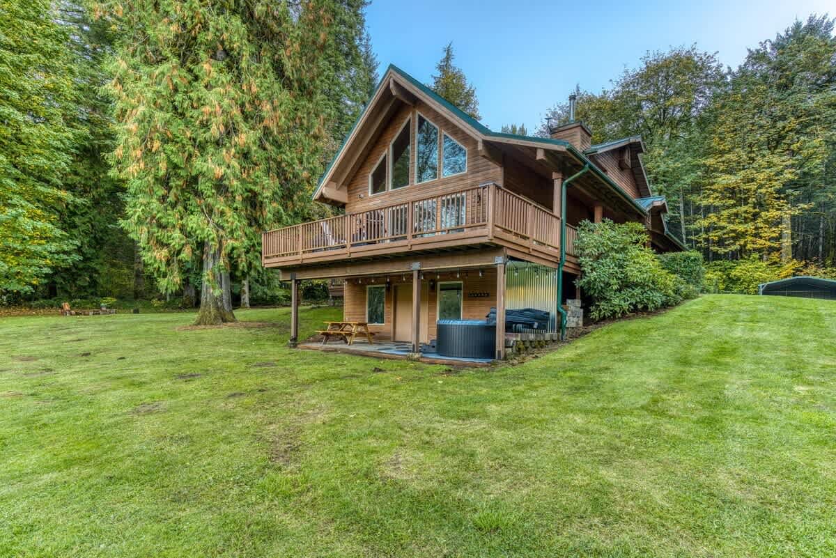 The side exterior of a multi-level wooden lodge featuring a large upper deck with a railing and a lower patio area with a hot tub and picnic table, surrounded by lush green grass and tall evergreen trees.
