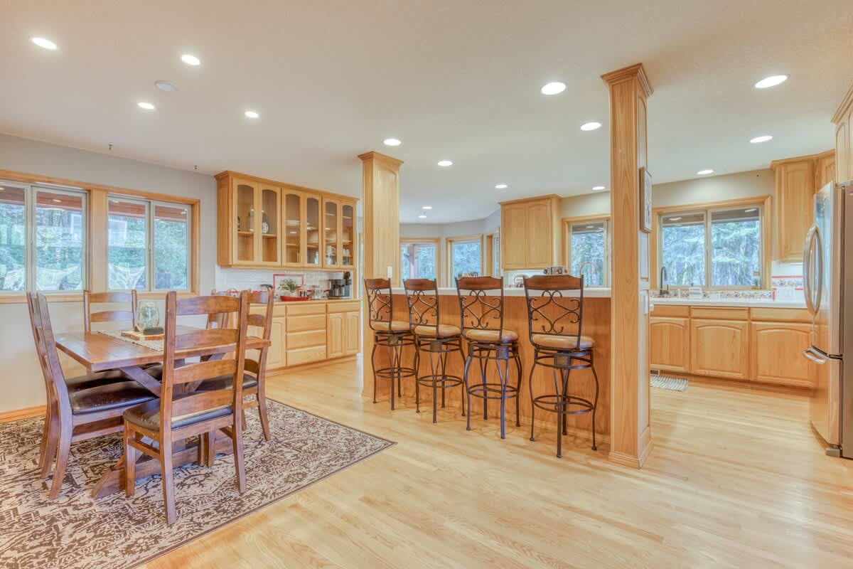 Expansive open-concept kitchen and dining space with light wood cabinetry, hardwood floors, and a large kitchen island. A wooden dining table with chairs sits on an area rug, and multiple windows provide natural light.