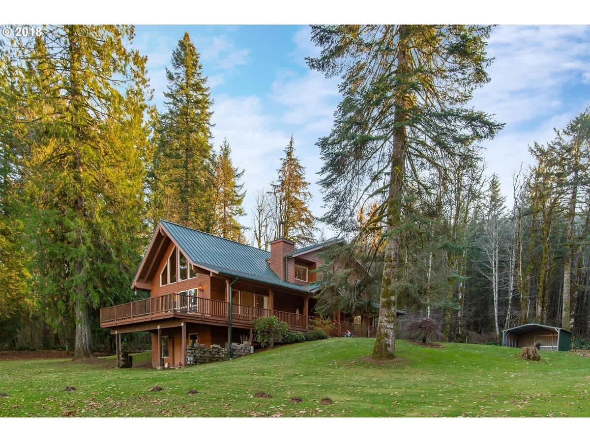 A large, rustic wooden house with a green metal roof is nestled on a grassy hill surrounded by tall evergreen trees under a bright blue sky.