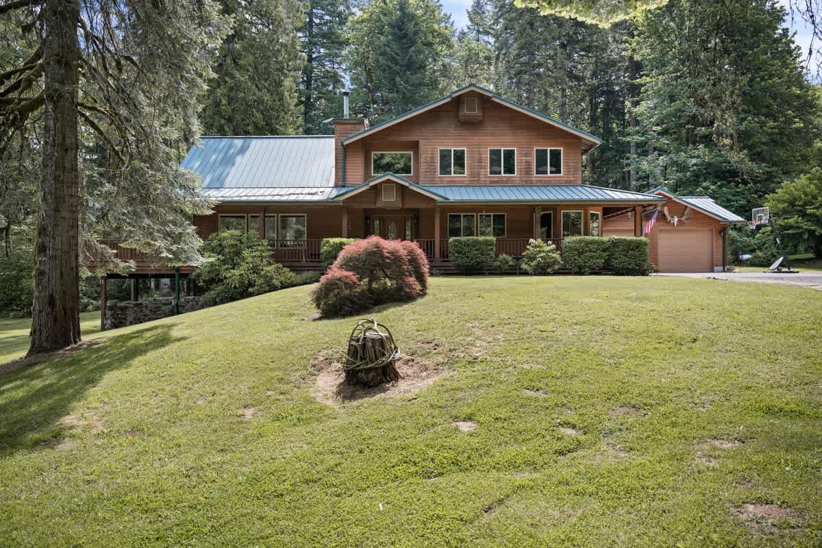 The front of a charming wooden house with a green metal roof is nestled on a grassy slope, surrounded by tall evergreen trees, with a paved driveway leading to the garage visible on the right.