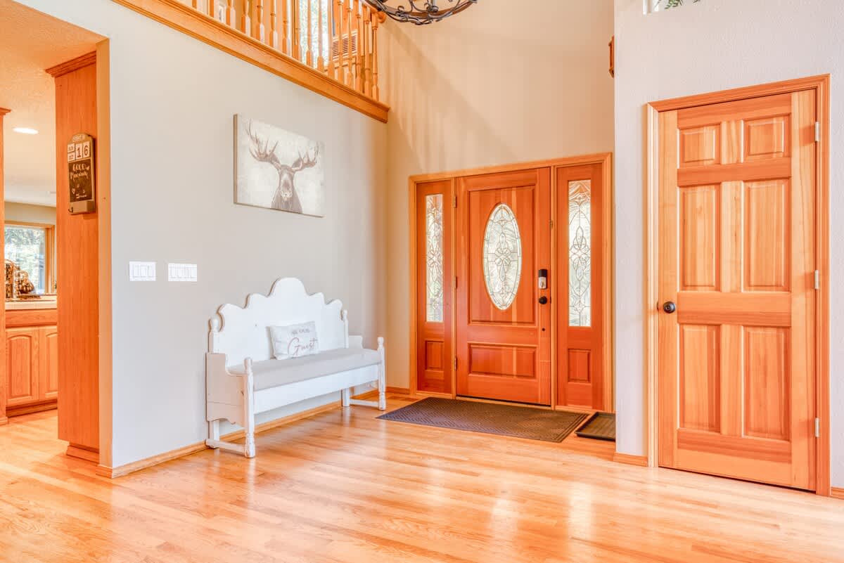 A bright foyer features polished hardwood floors, a light wood front door with glass inserts, a white decorative bench, and a large deer art print on the wall.