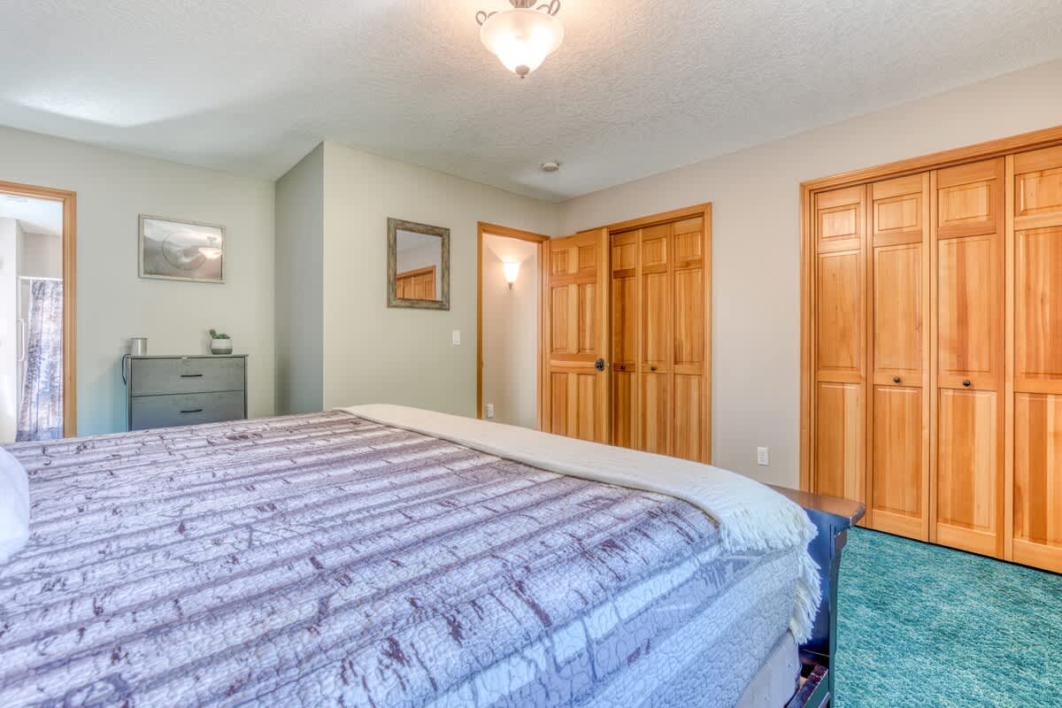 View of a spacious bedroom with a large bed, showing light wood bi-fold closet doors and a dresser. A doorway leads to an adjacent bathroom, and the room features teal carpet.