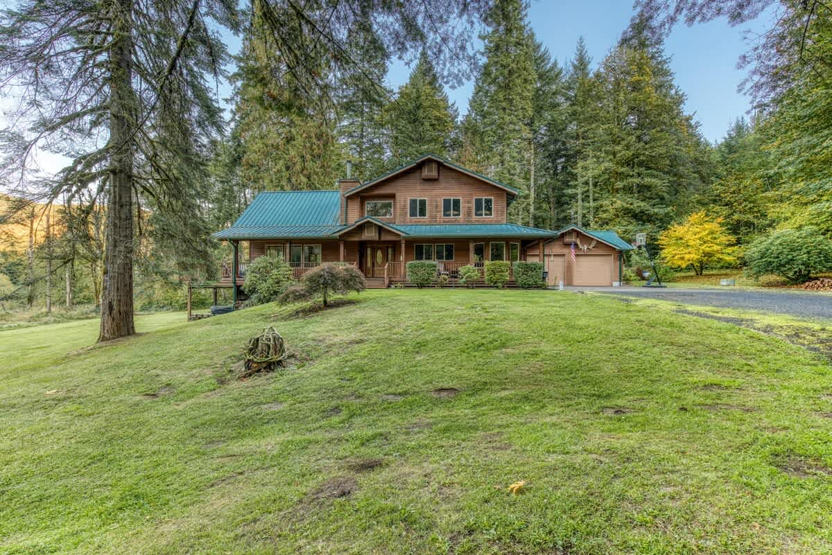 The front exterior of a large, rustic wooden lodge with a green metal roof, multiple windows, and an attached garage, nestled among tall evergreen trees on a grassy slope.
