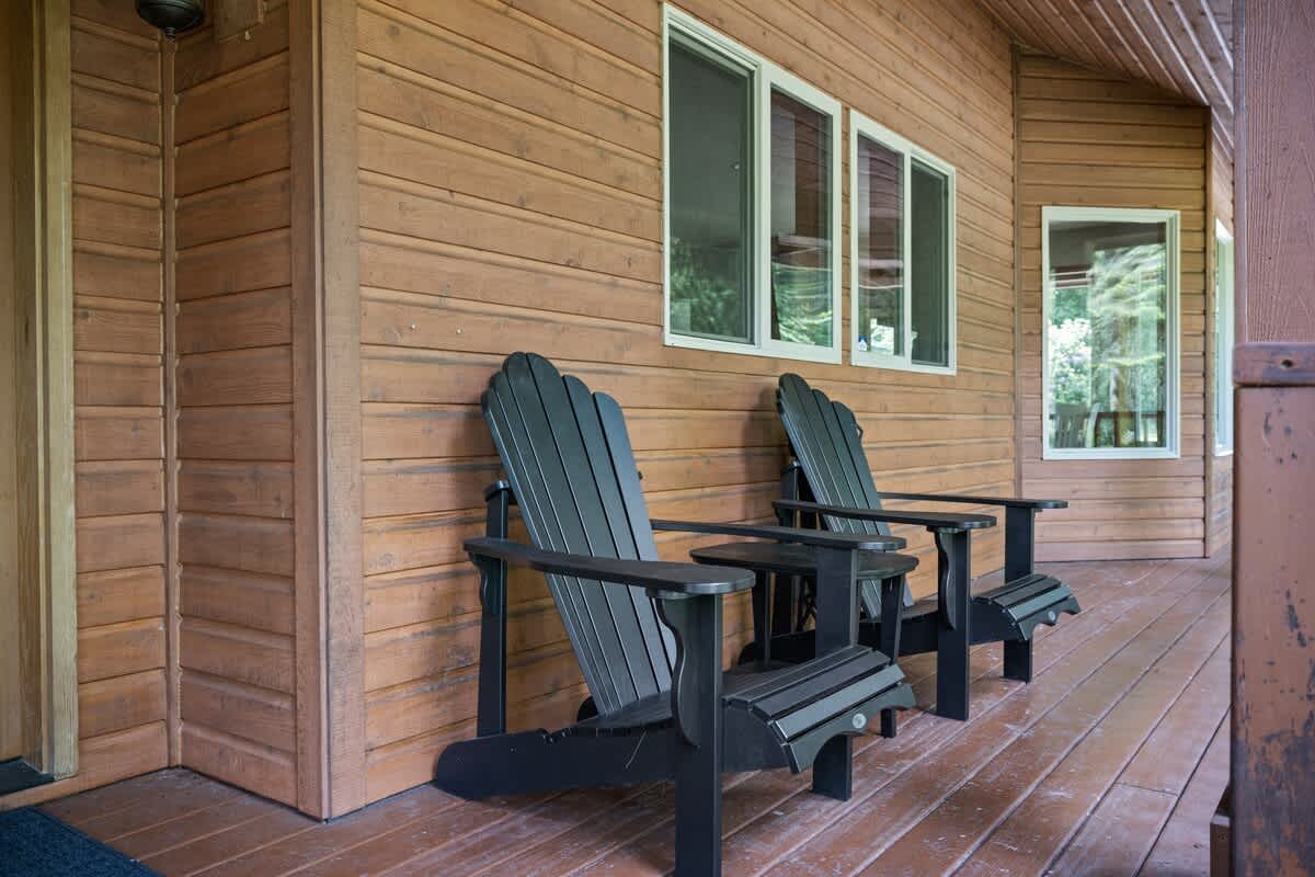Two black Adirondack chairs and a small side table are placed on a wooden deck next to the rustic wooden siding of the house, with large windows reflecting the surrounding greenery.