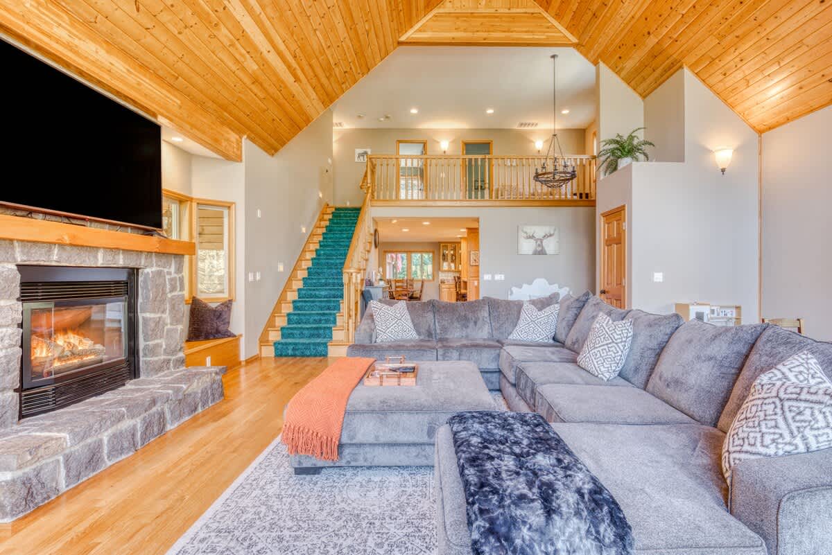 A cozy living room with a large gray sectional sofa, a stone fireplace with a TV mounted above, and a teal carpeted staircase leading to an upper level. The room has light hardwood floors and a vaulted wooden ceiling.