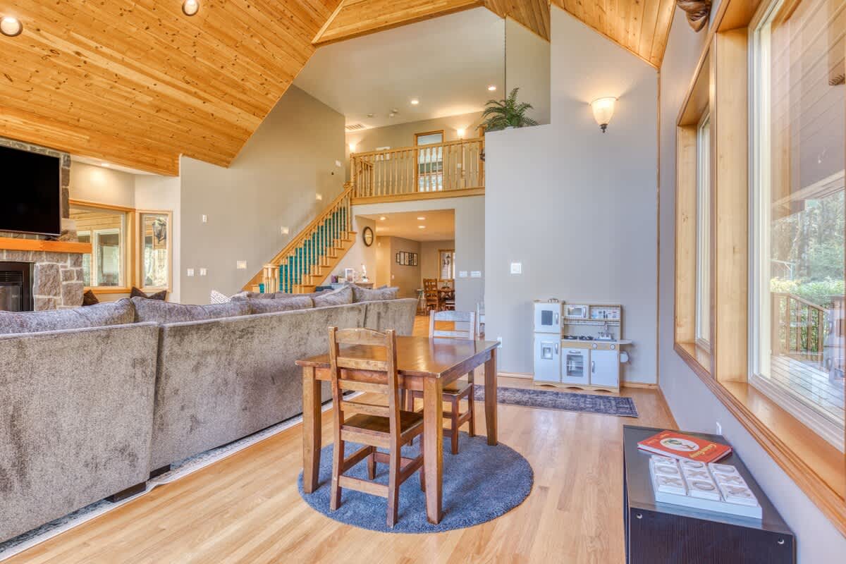 A bright living space with a small wooden dining table and chairs, a children's play kitchen, and a view towards a large gray sectional sofa. The room features light hardwood floors and a vaulted wooden ceiling, with a mezzanine visible in the background.