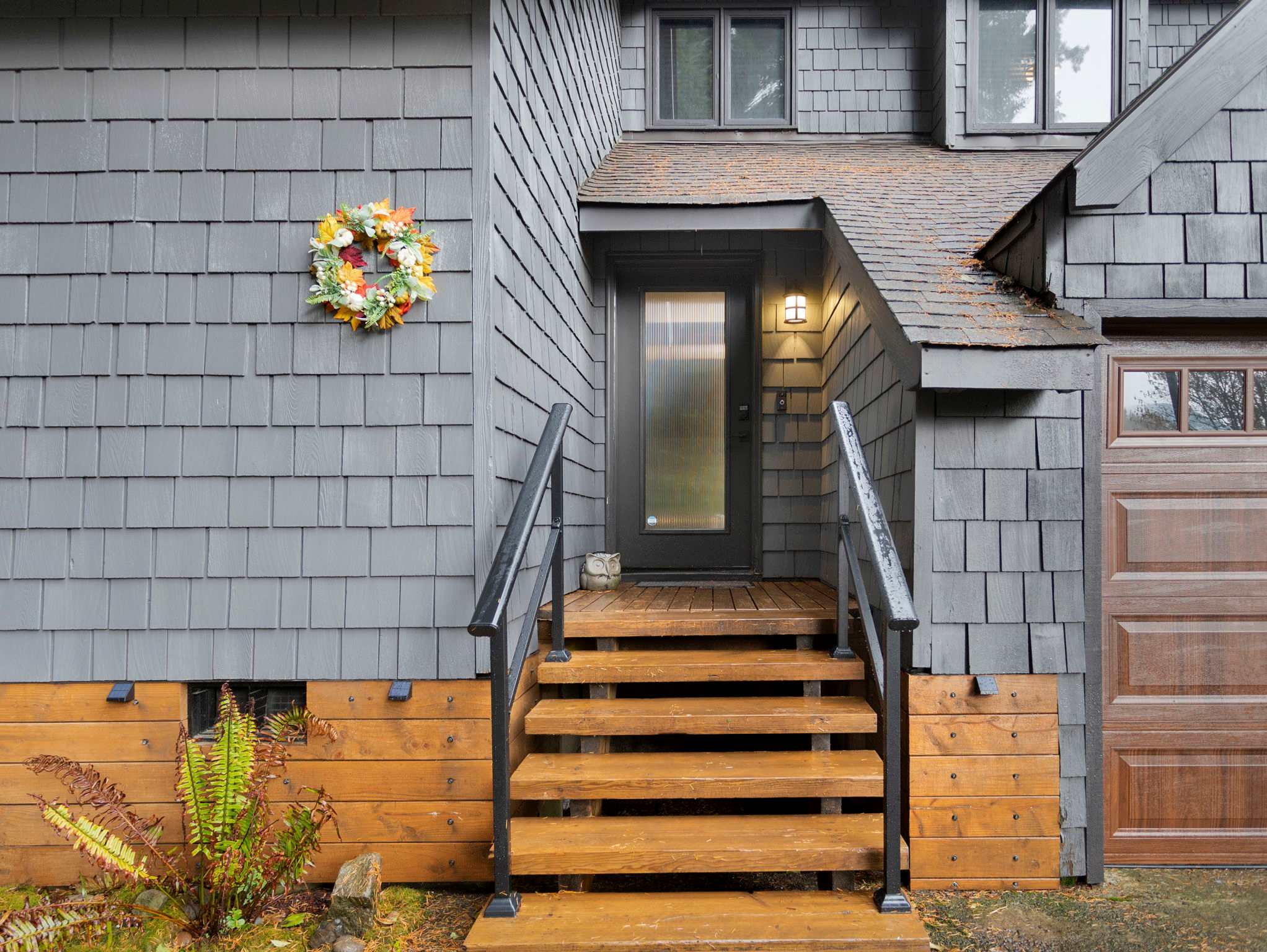 The front entrance of the house, featuring wooden steps with black railings leading to a dark front door. A fall-themed wreath hangs on the dark gray shingle siding.