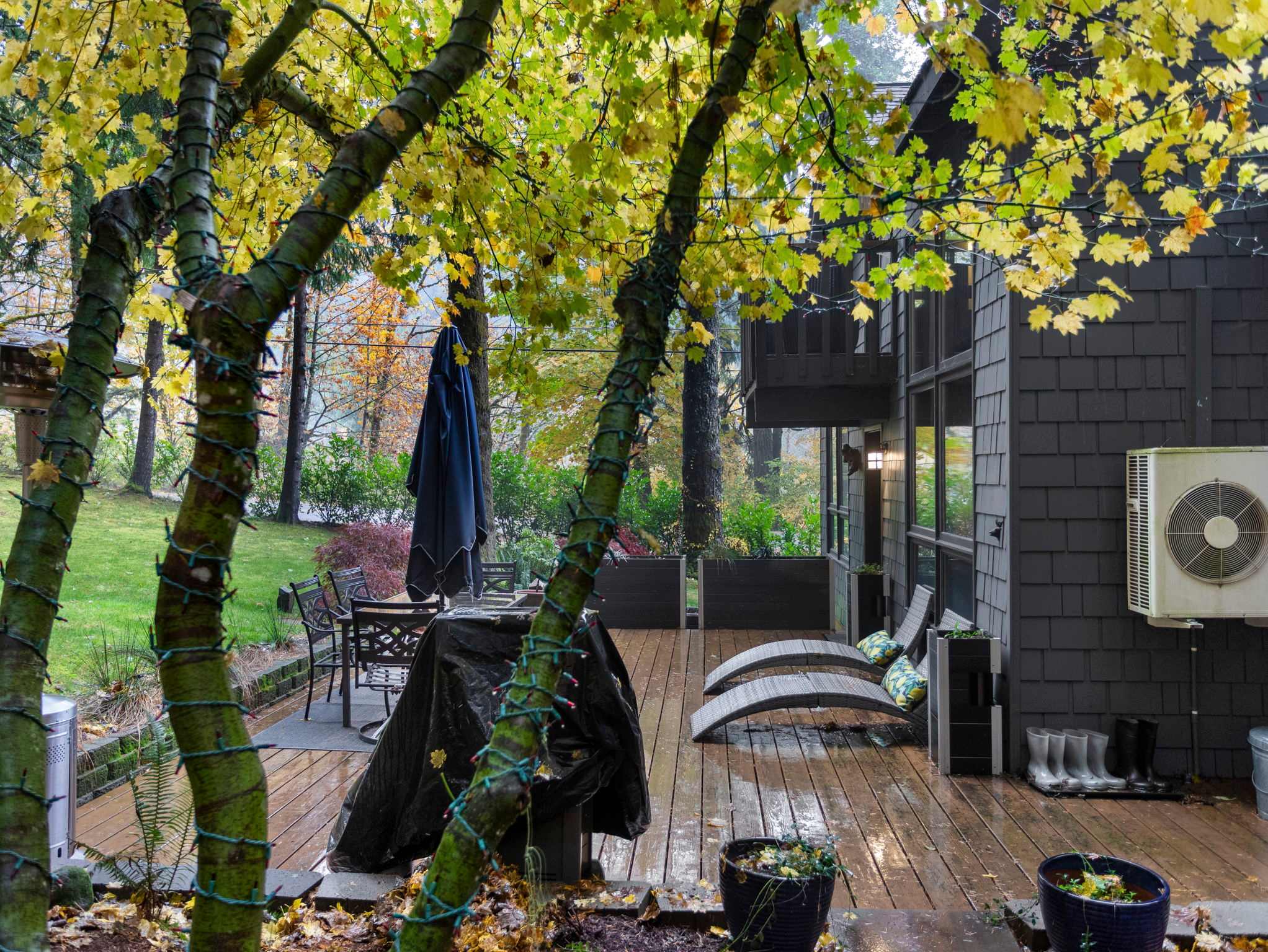A wooden deck features an outdoor dining set with a dark umbrella and two lounge chairs, surrounded by vibrant green and yellow-leafed trees. The dark gray house exterior with an air conditioning unit is visible to the right.