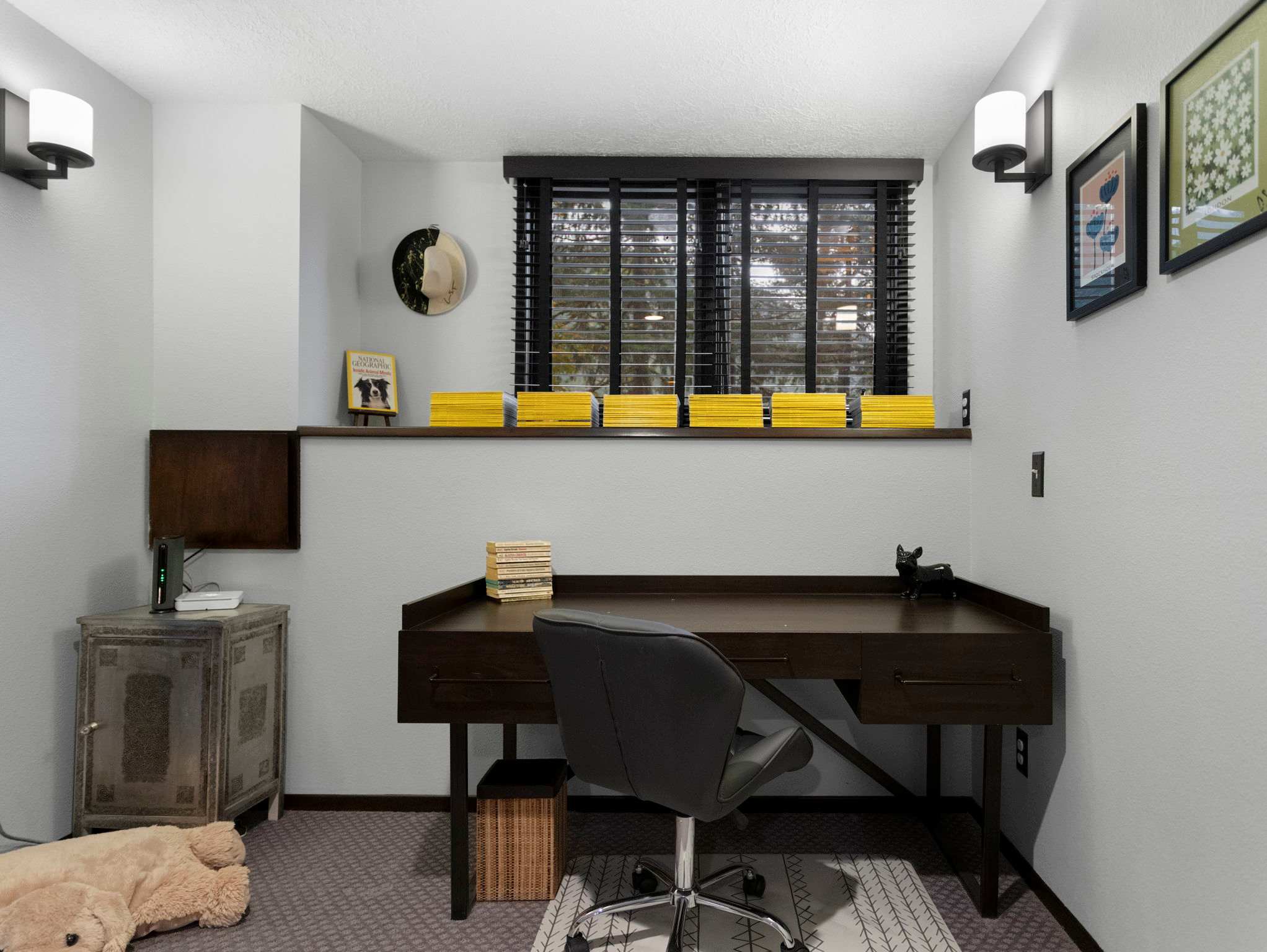 A detailed view of the dark wood desk area, featuring an office chair, a window with dark blinds, and a small rustic side table. Books and decorative items are neatly arranged on the desk and a shelf above the window.