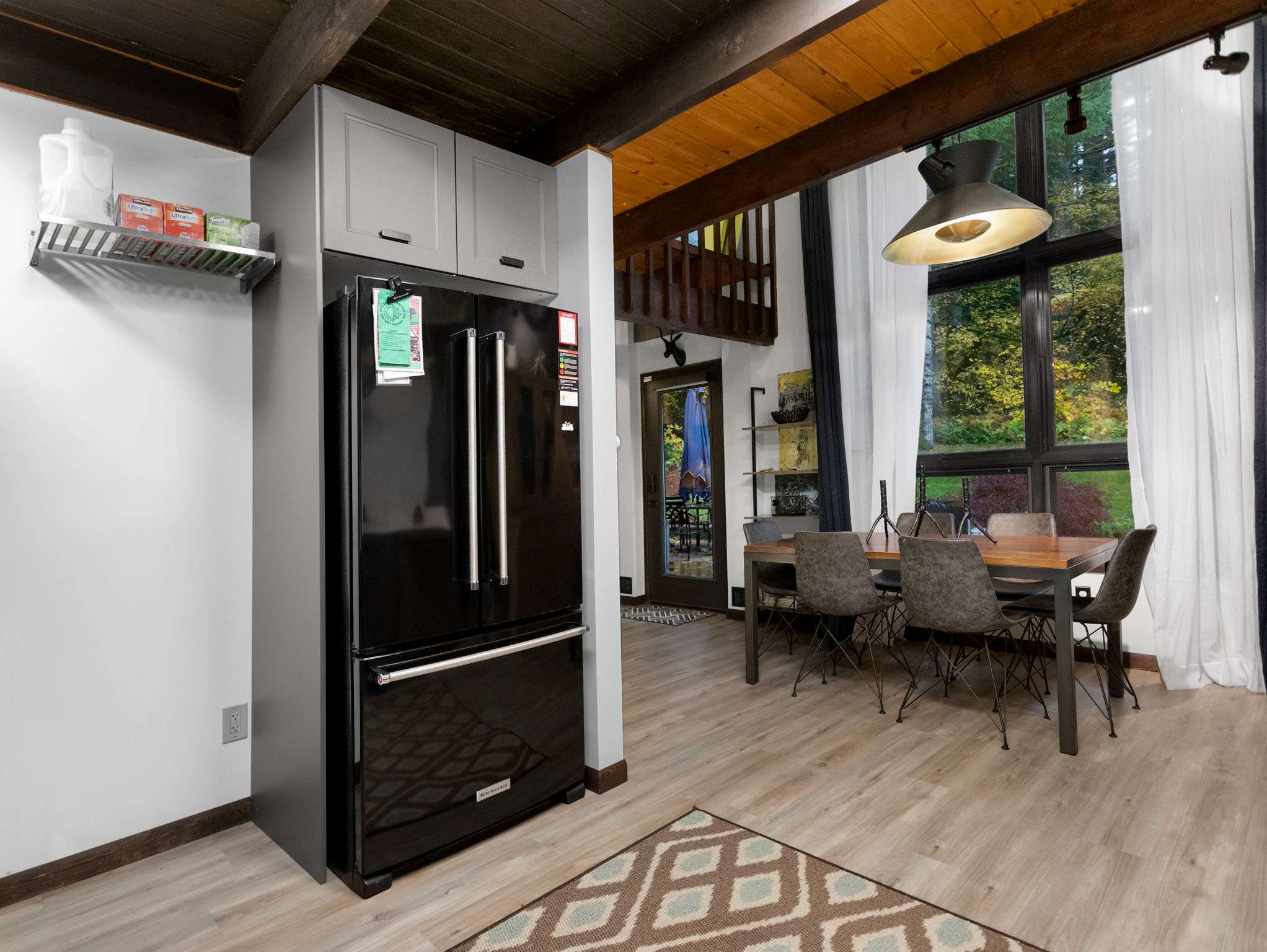 A spacious open-plan area showcasing a black French-door refrigerator integrated into light grey cabinetry, adjacent to the dining table. Beyond, large windows offer a view of the outdoors, and the kitchen area is visible in the background.