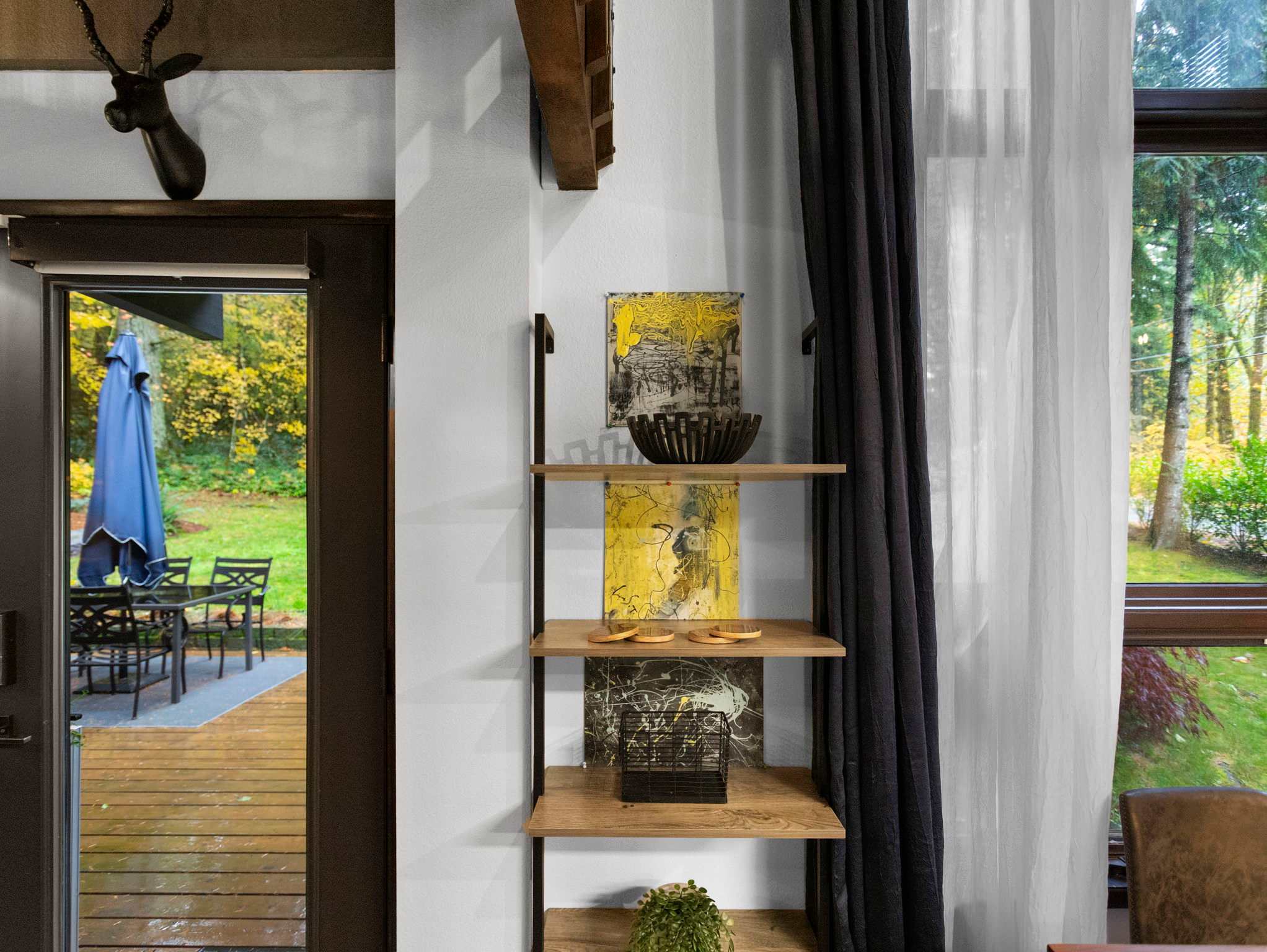 Close-up of a wooden deck with an outdoor dining set and a blue umbrella, visible through a glass door. A tall shelving unit with decorative items is inside the house, next to the door.