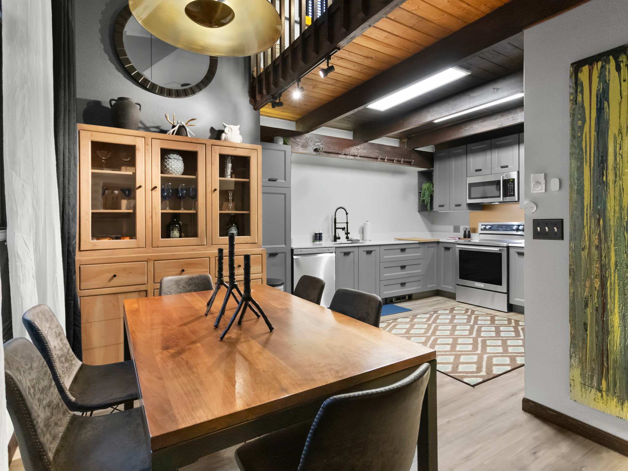An open-concept dining and kitchen area features a large wooden dining table with grey chairs, under a striking gold pendant light. The kitchen has light grey cabinetry, stainless steel appliances, and a patterned rug, all beneath exposed wooden beams.