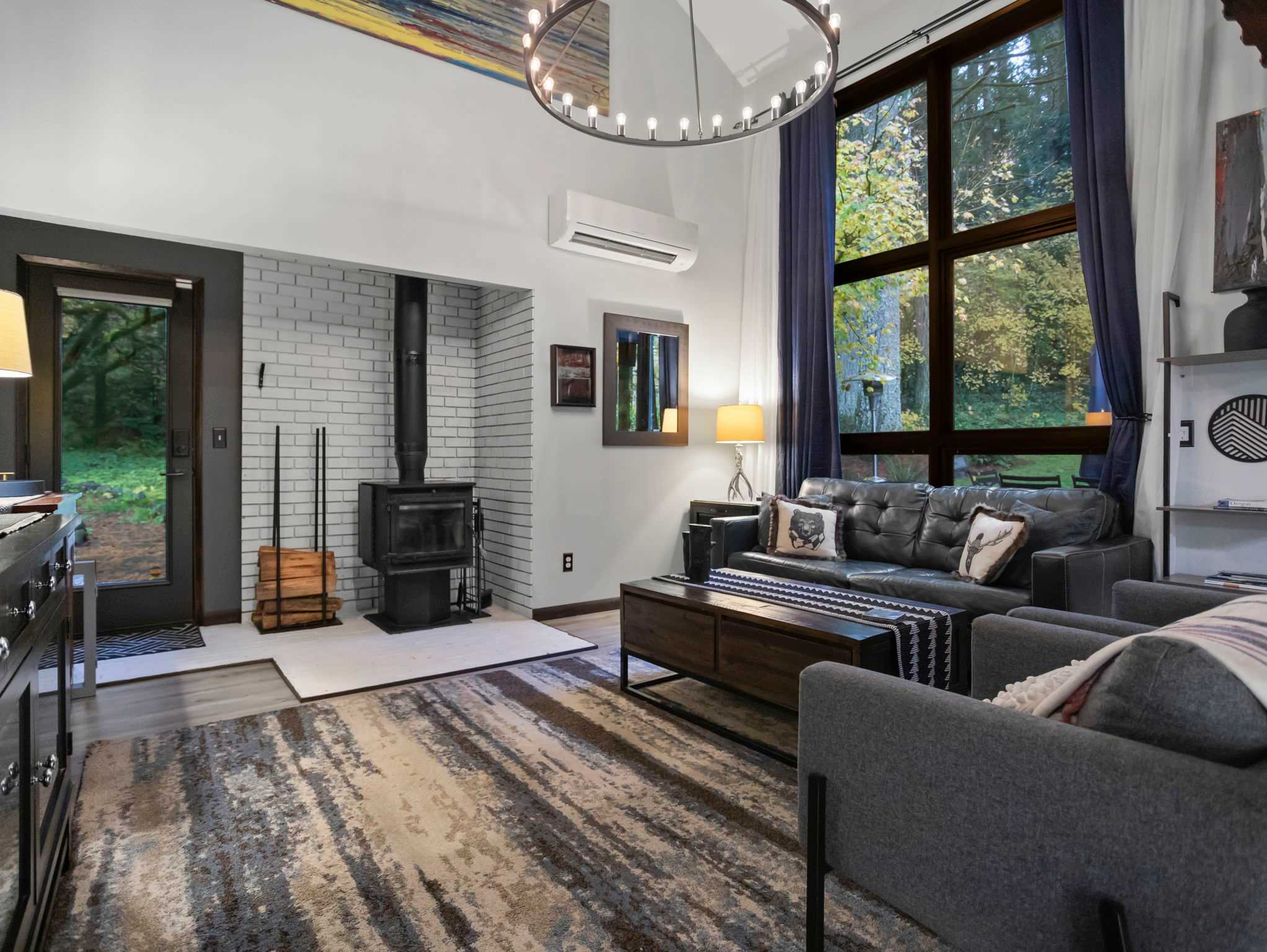 Cozy living room featuring a black leather sofa, a wood-burning stove set against a white brick wall, and a large window overlooking a lush forest. A patterned rug covers the light wood floor, and a modern chandelier hangs from the ceiling.