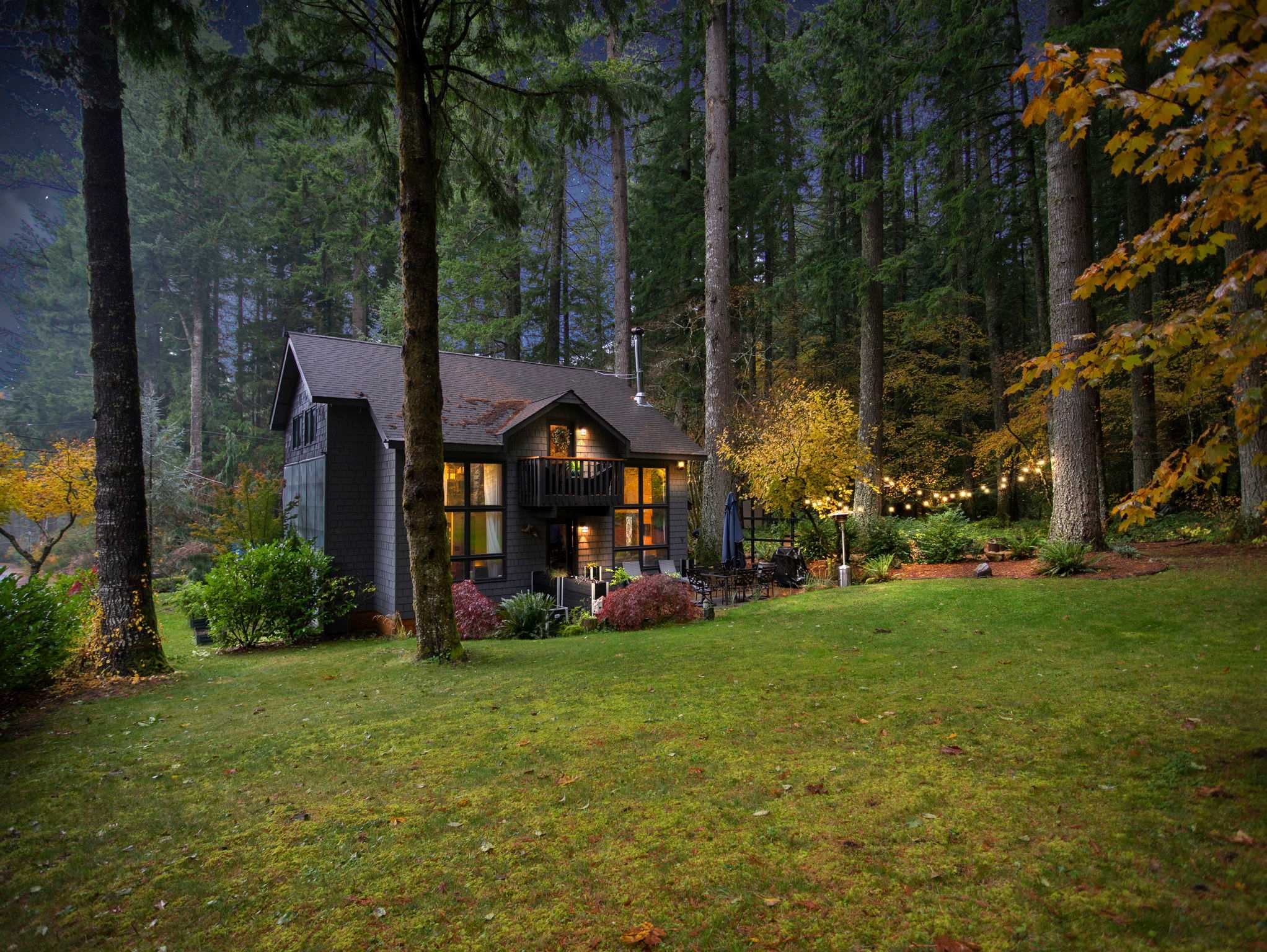 Charming dark-sided house nestled among tall evergreen trees and vibrant autumn foliage, illuminated by string lights and exterior lighting under a twilight sky. A grassy yard and outdoor seating are visible.