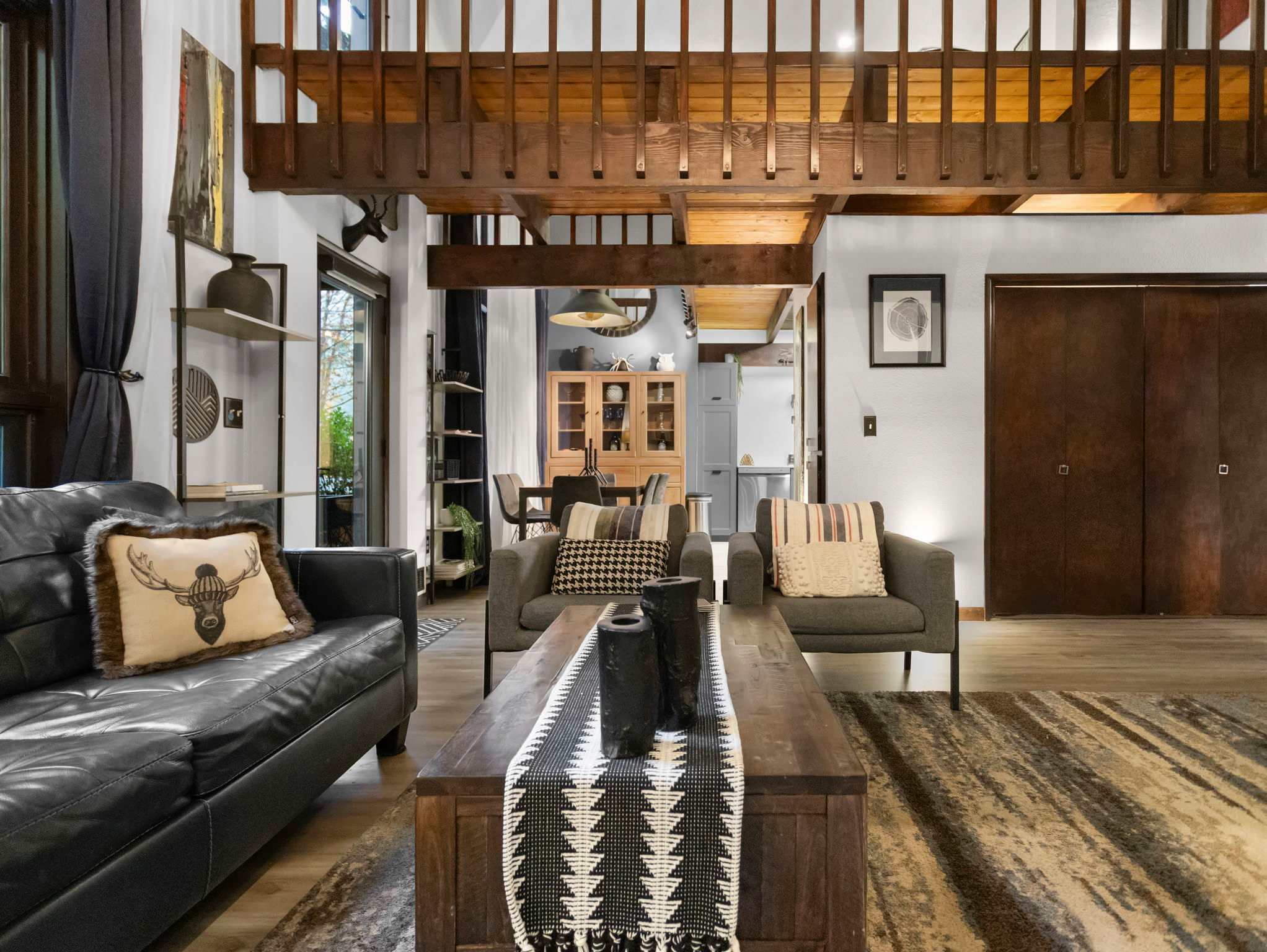 View of the living room looking towards the dining area and kitchen, featuring a black leather sofa, two gray armchairs, and a dark wood coffee table. A wooden railing from an upper level overlooks the space, and a large patterned rug anchors the seating area.