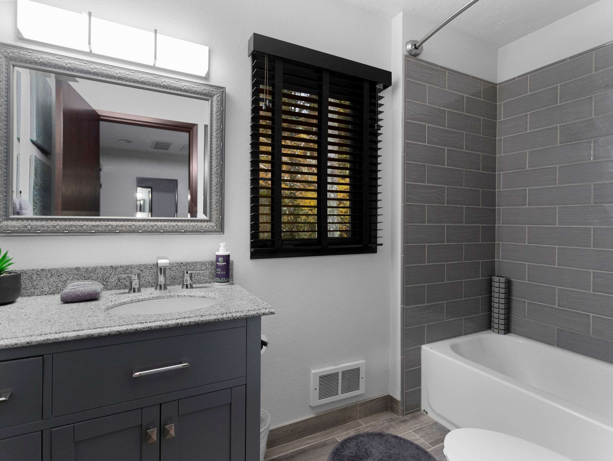 A modern bathroom featuring a grey vanity with a granite countertop and a white sink, beneath a large framed mirror. A bathtub with a shower is enclosed by grey subway tiles, and a window with dark blinds provides natural light.