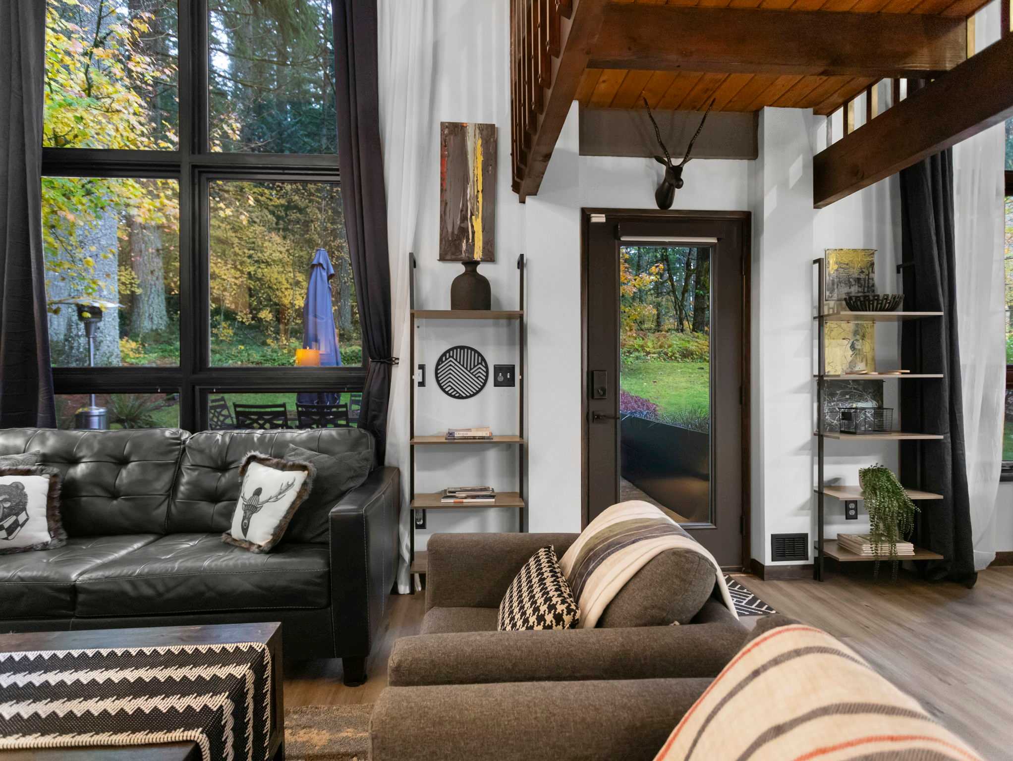 Corner view of the living room showing a gray armchair, a black leather sofa, and a door leading to an outdoor deck with a blue umbrella. A tall, dark wood shelving unit with decorative items stands against the wall.
