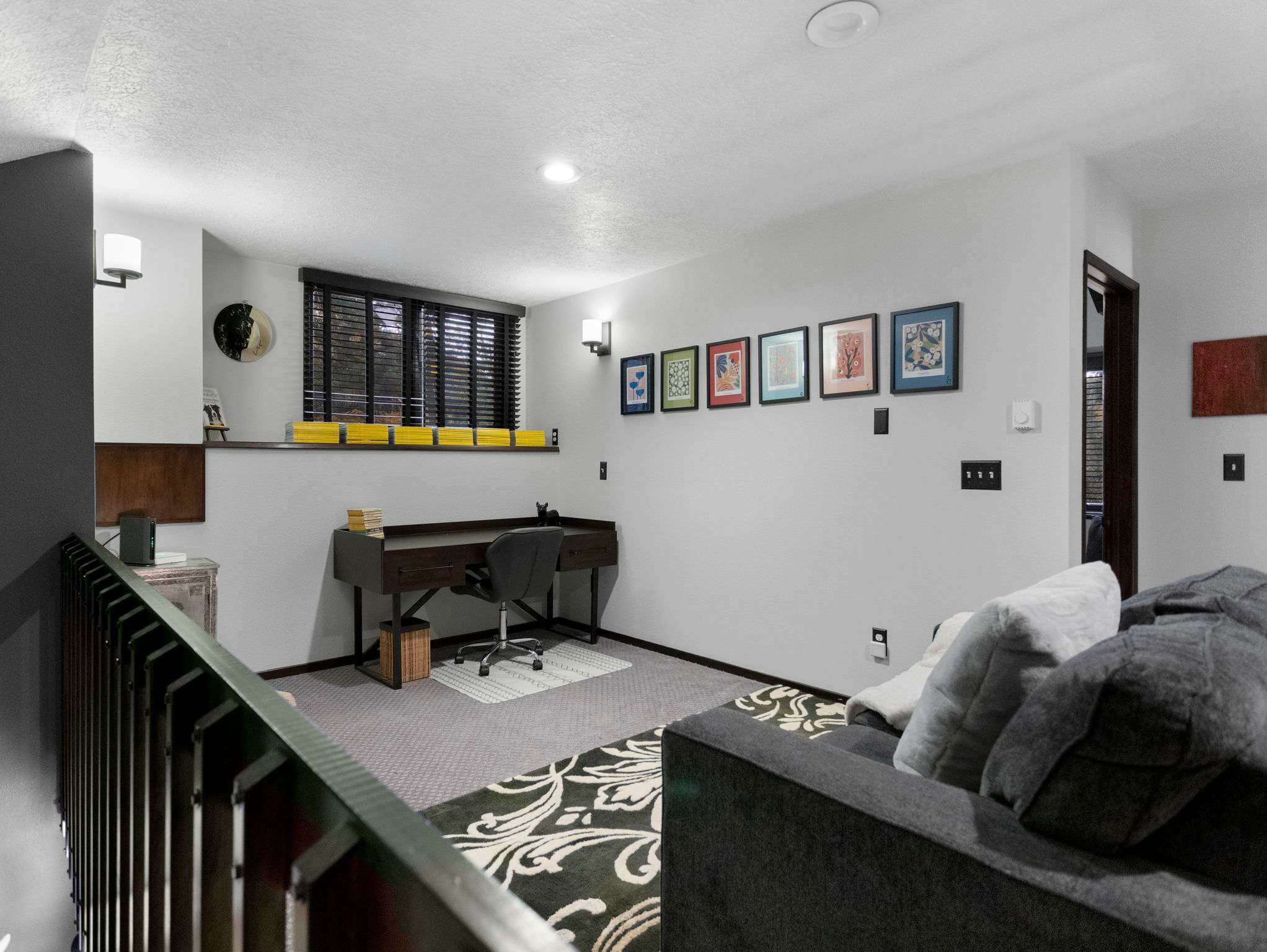 A bright loft space features a dark wood desk with an office chair, a grey sofa with pillows, and a window with dark blinds. The light walls are adorned with a series of colorful framed artworks, and a railing is visible in the foreground.