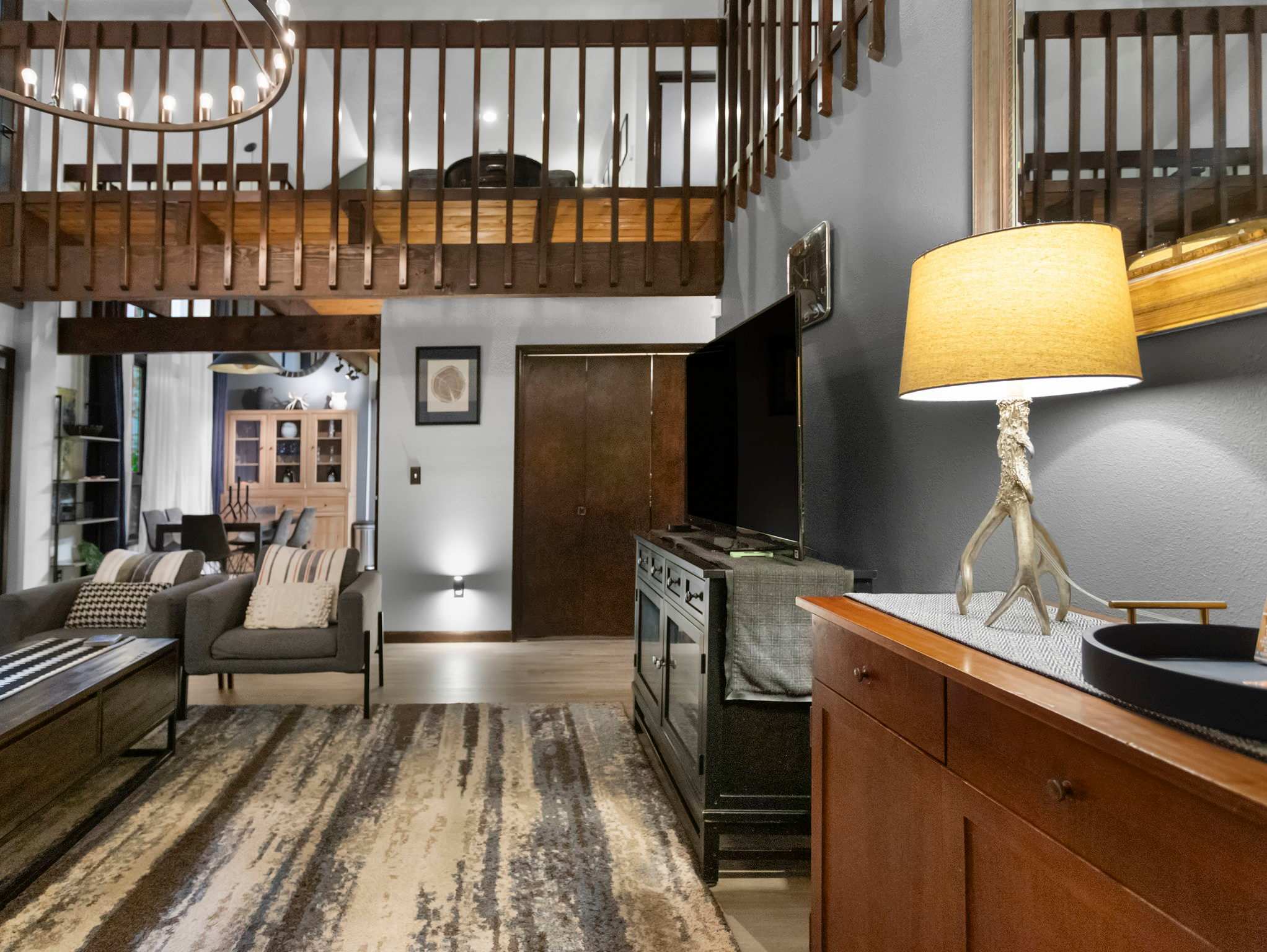 Living room view showing a TV on a dark media console, a wooden side table with a lamp, and a large patterned rug. The open layout reveals a glimpse of the dining area and kitchen beyond, with a wooden railing from an upper floor.
