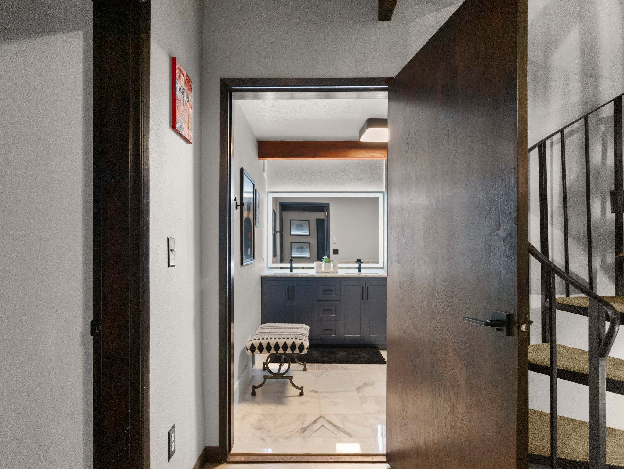 A dark wooden door stands ajar, revealing a modern bathroom with a double vanity featuring a large illuminated mirror and dark grey cabinetry. White marble-look tile flooring and a patterned bench are visible inside.