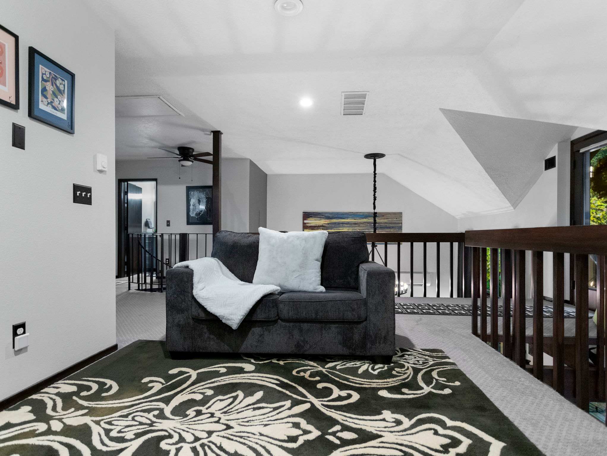 A cozy grey sofa with a light blue throw and pillow sits on a dark green and white patterned rug in an open loft area. A dark wood railing separates the space, with a ceiling fan visible in the background.