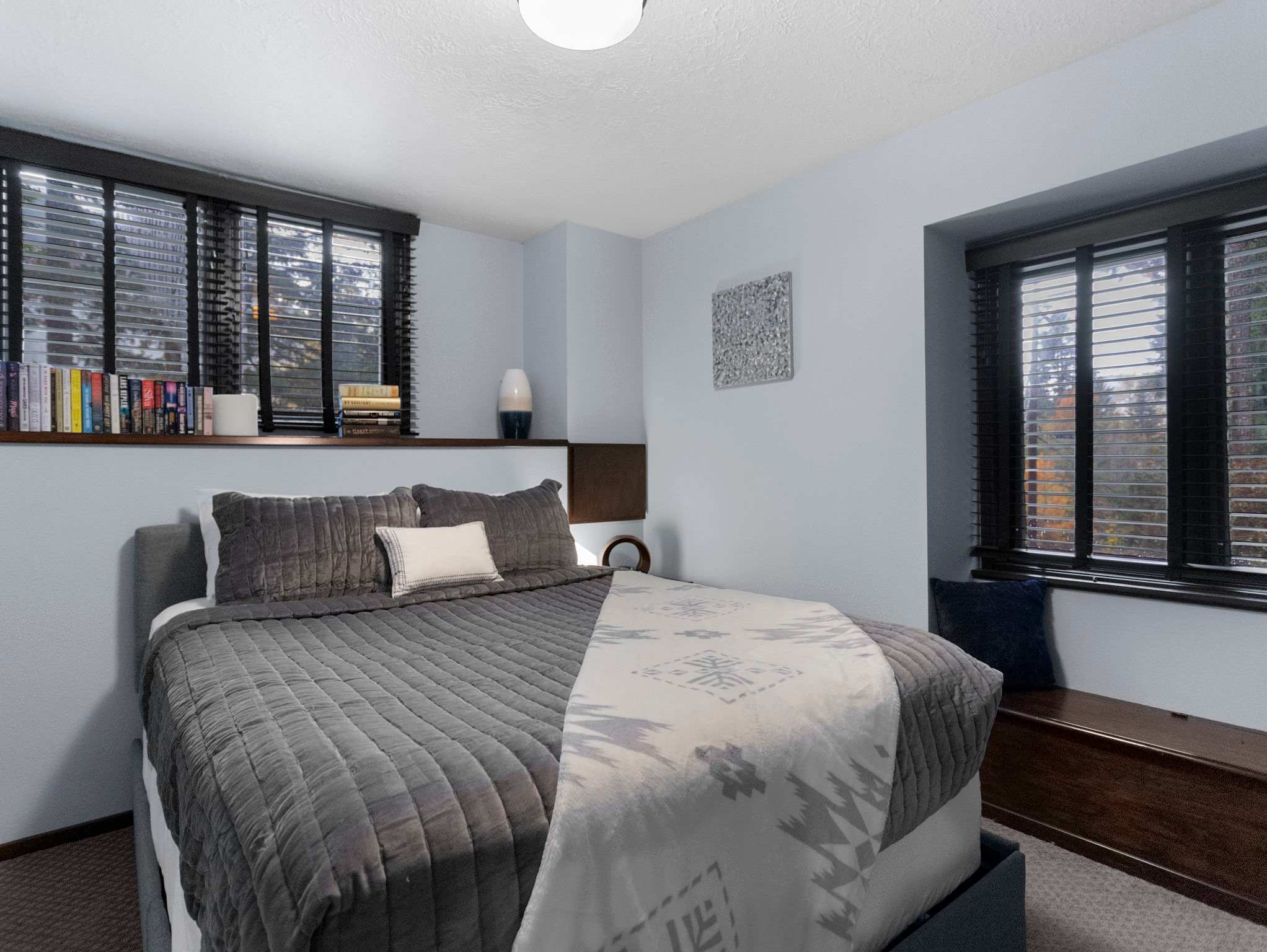 A serene bedroom with a large bed dressed in grey bedding and a white patterned throw, flanked by two windows with dark wood blinds. A wooden bench sits beneath one window, and a shelf above the other holds books and a decorative vase.
