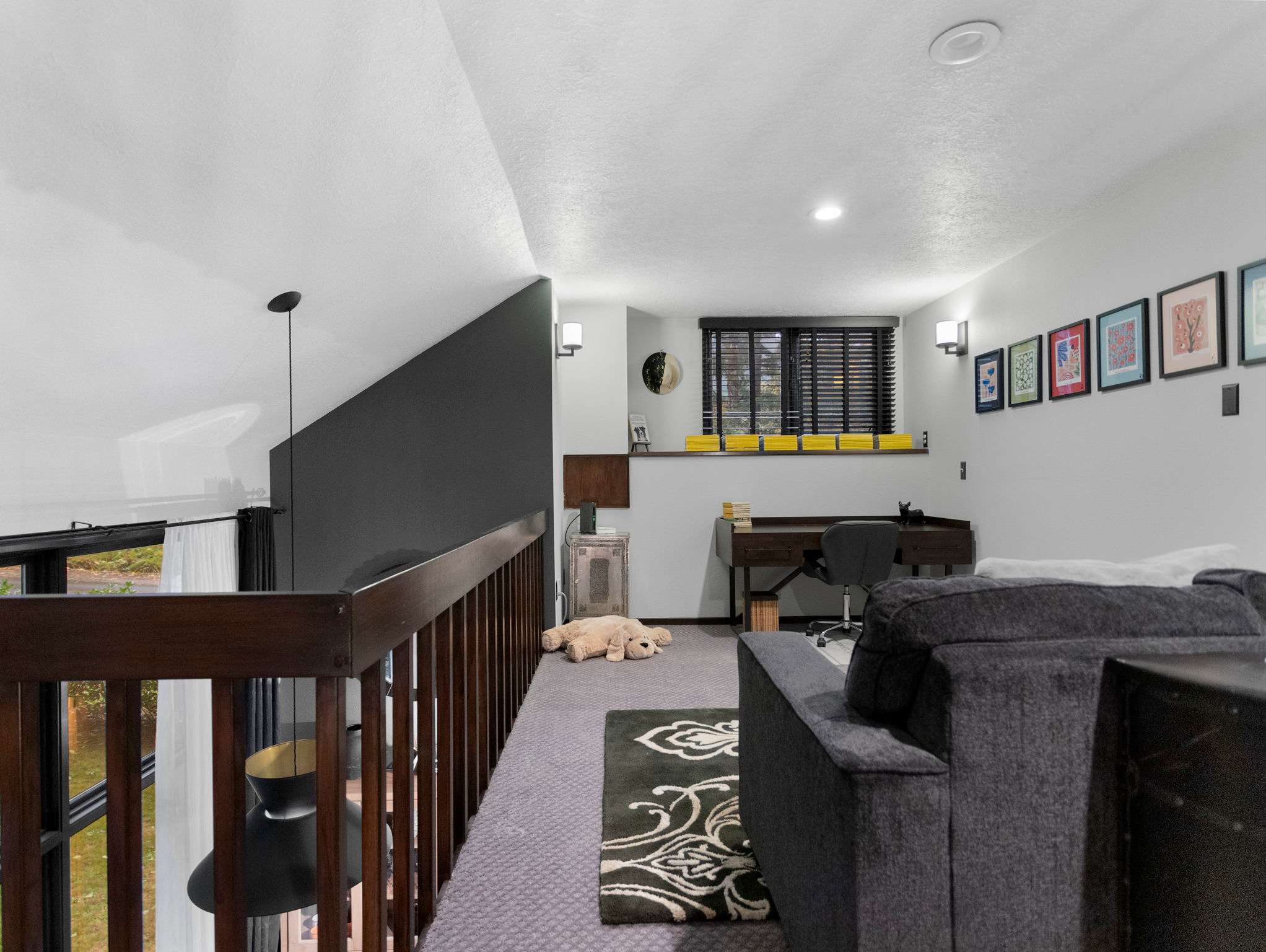 Another view of the loft area, configured as a lounge or office space with a grey sofa, a desk, and a chair. The room features a dark accent wall, a window with blinds, and framed artwork.
