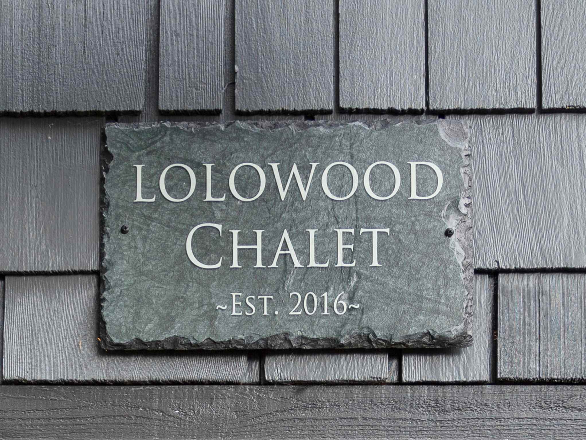 A close-up of a slate sign on the dark gray shingle siding of the house, engraved with "LOLOWOOD CHALET ~EST. 2016~".