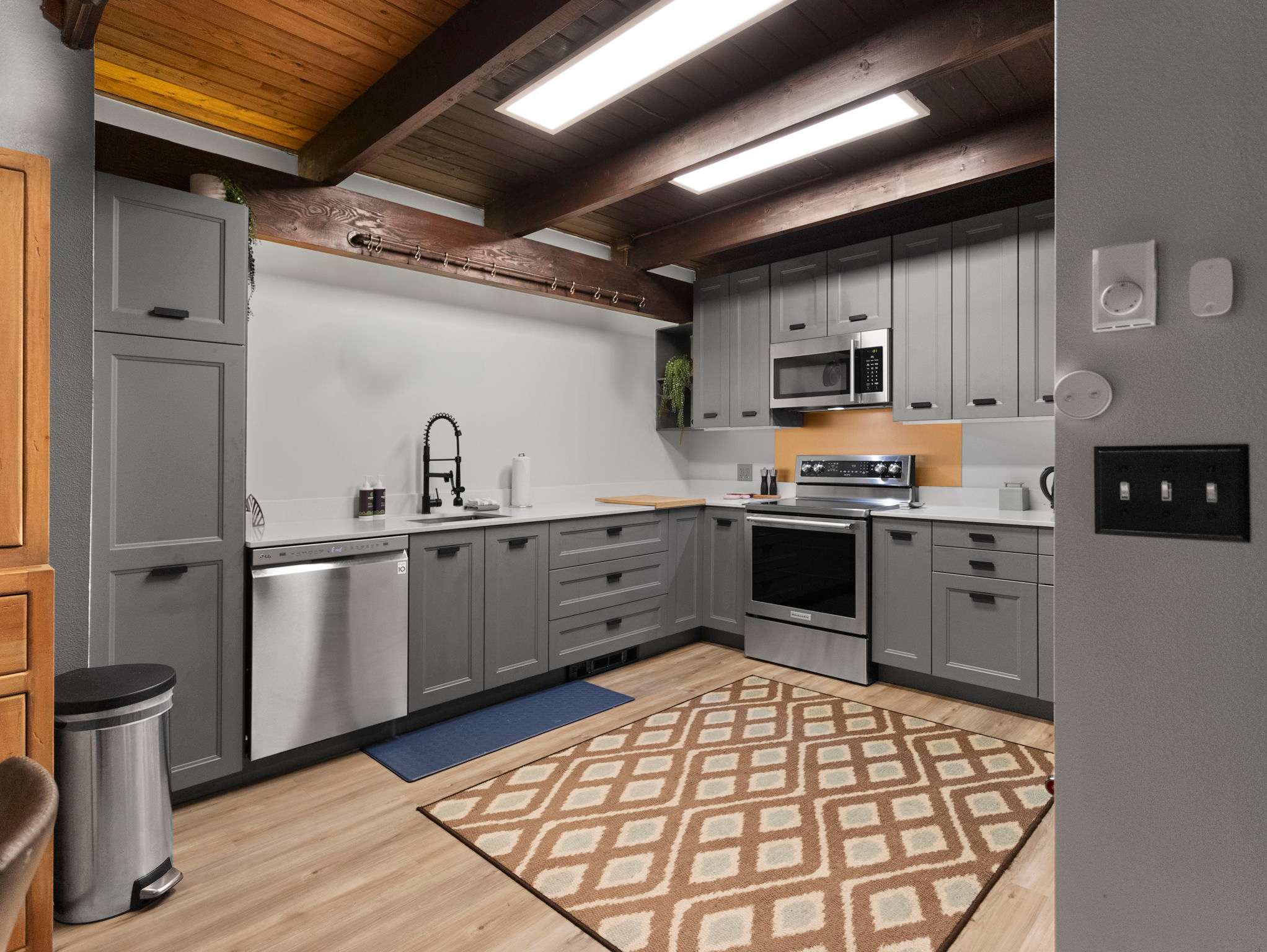 A modern kitchen with light grey shaker-style cabinets, white countertops, and stainless steel appliances including a dishwasher, range, and microwave. A brown and cream geometric patterned rug covers part of the light wood-look flooring, under exposed wooden beams.