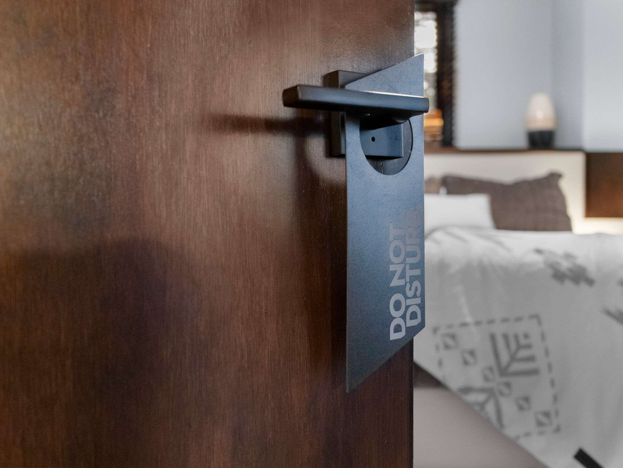 A close-up of a dark wood door handle with a black "DO NOT DISTURB" sign hanging from it, offering a soft-focus glimpse of a bed and bedroom beyond.