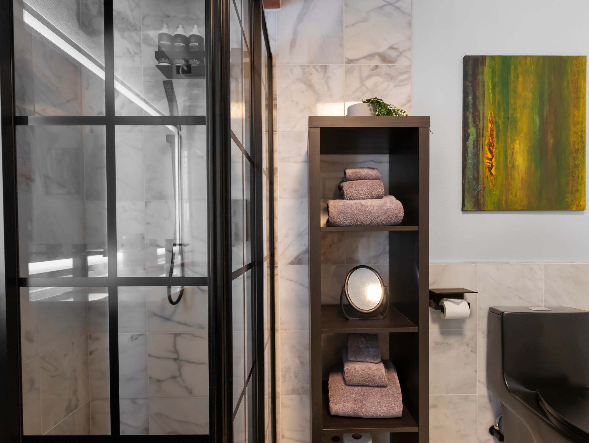 A modern bathroom features a walk-in shower with black-framed glass doors and white marble-look tiles, alongside a dark wooden shelving unit holding folded towels and a small mirror. A dark toilet is partially visible next to the shelving.